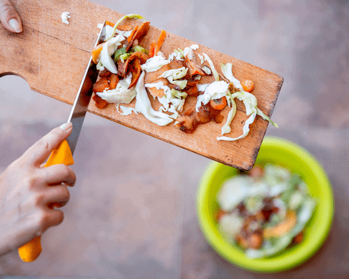 The image shows a person's hand using a knife to slide veggie waste off the board. Below the board, there's a green bowl with compost. 