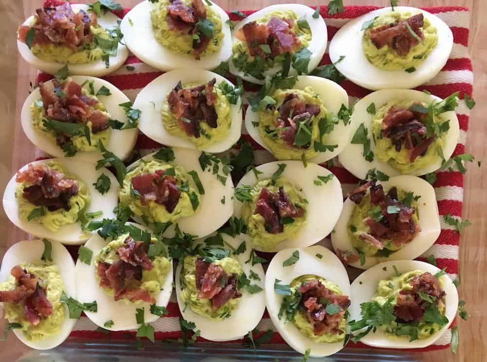 A platter of avocado deviled eggs for camping topped with bacon and parsley, arranged on a red and white striped cloth, a delicious twist on a classic appetizer ready to be served.