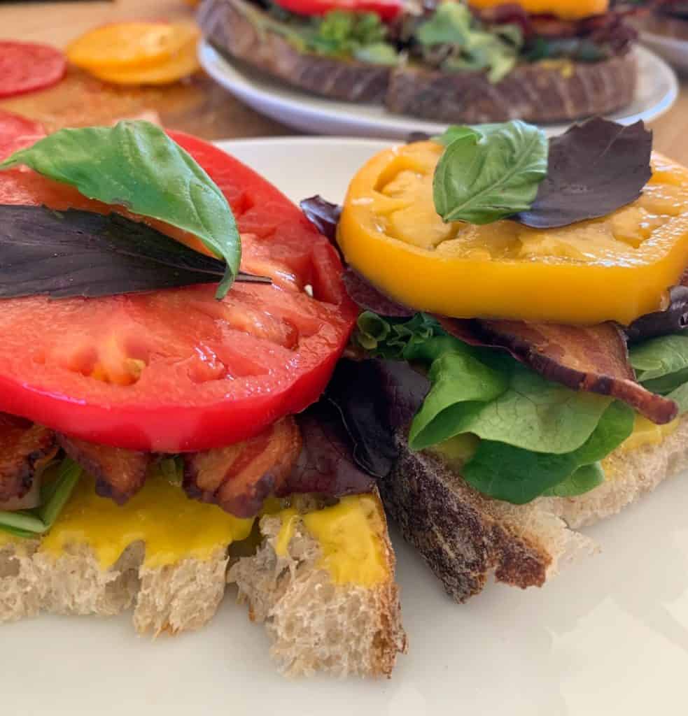 A close up of a summer-kissed open-face BLT with fresh sliced yellow tomatoes, garden lettuce and basil, aioli, and thick-sliced bacon on crusty bread.