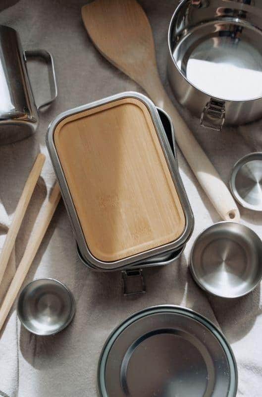 Assortment of camping kitchenware neatly arranged on a cloth, including a stainless steel pot, a bamboo-covered container, and various cooking utensils, all ready for an eco-friendly outdoor cooking experience.