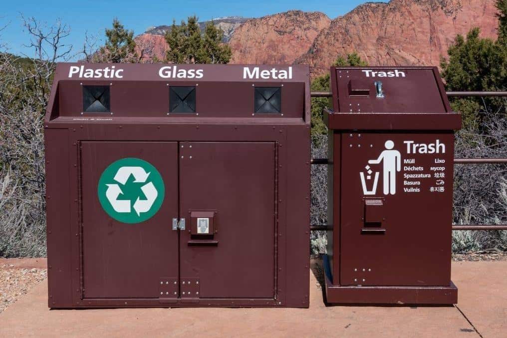 A recycling station with separate compartments for "Plastic," "Glass," and "Metal," indicated by labels above each slot. Adjacent to this is a trash bin labeled "Trash" with corresponding waste disposal icons and the word "trash" written in various languages. The bins are set against a natural backdrop with trees and red rock formations.