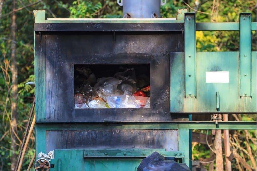 An overfilled green industrial waste incinerator with a pile of garbage bags waiting to be burned, highlighting issues of waste management and environmental impact.
