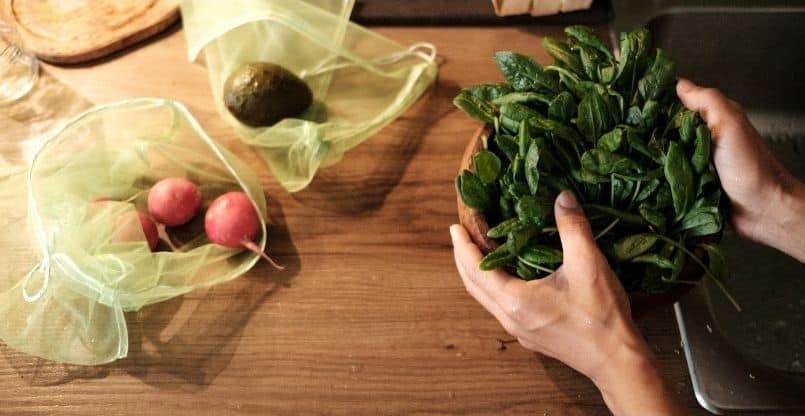 Hands washing fresh spinach in a wooden bowl beside a kitchen sink, with reusable mesh bags containing radishes and an avocado on a wood countertop, promoting eco-friendly food preparation.