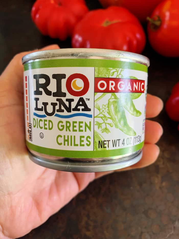 My hand holds a can of Rio Luna diced green chiles which is an ingredient for the casserole. Red tomatoes are seen in the background.