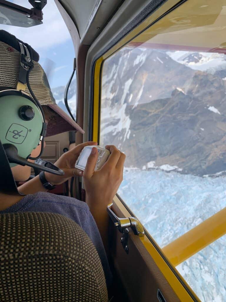 View from inside a small aircraft with a passenger wearing a headset, holding a camera, capturing the stunning aerial view of a glacier and rugged mountain terrain below.