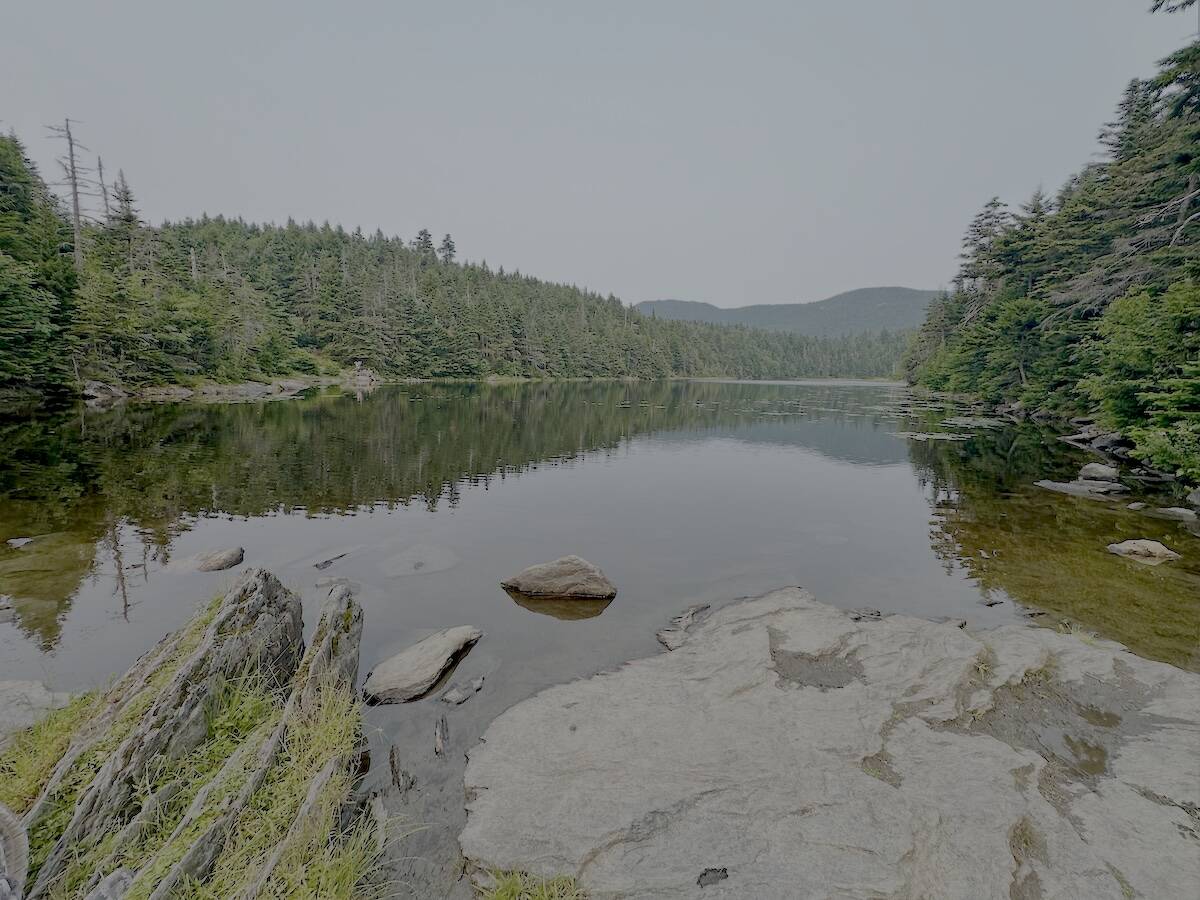 A calm mountain lake bordered by dense evergreen forest under a hazy sky, with large rocks in the foreground leading into the clear, reflective water. The stillness of the lake mirrors the trees and distant hills, creating a serene, untouched wilderness scene.