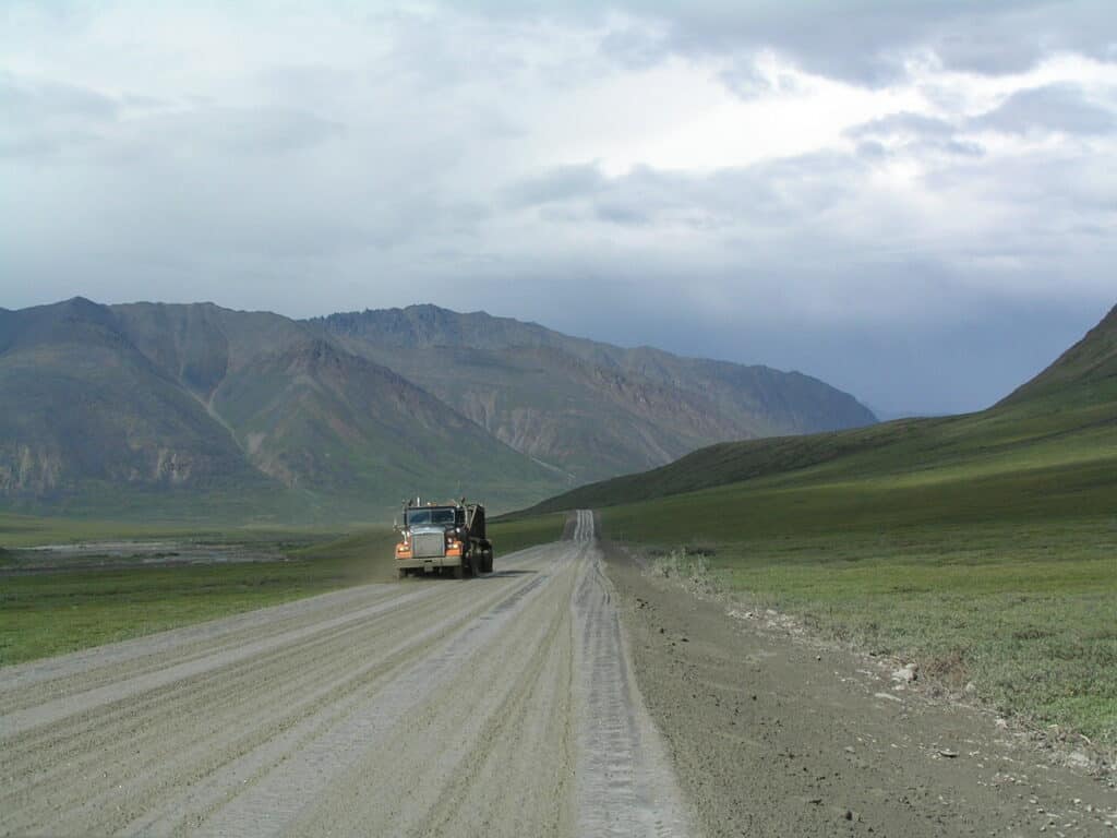The Dalton Highway has approximately 250 trucks travel on it daily! This shows the gravel highway that goes on and on into and past the huge mountains. A large truck approaches on the highway.