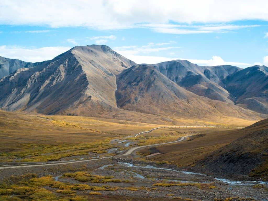 Panoramic view of the Dalton Highway in Alaska, winding through a vast valley with golden autumn tundra leading up to rugged mountain peaks. The road serves as a human-made ribbon through the wild and untouched landscape, under a soft blue sky with wispy clouds.
