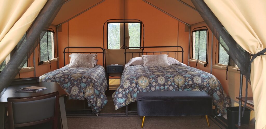Two beds, one twin and one double, are set up inside a warm tan canvas wall tent with windows. The bedding is floral with blues and there is a side table with two chairs.