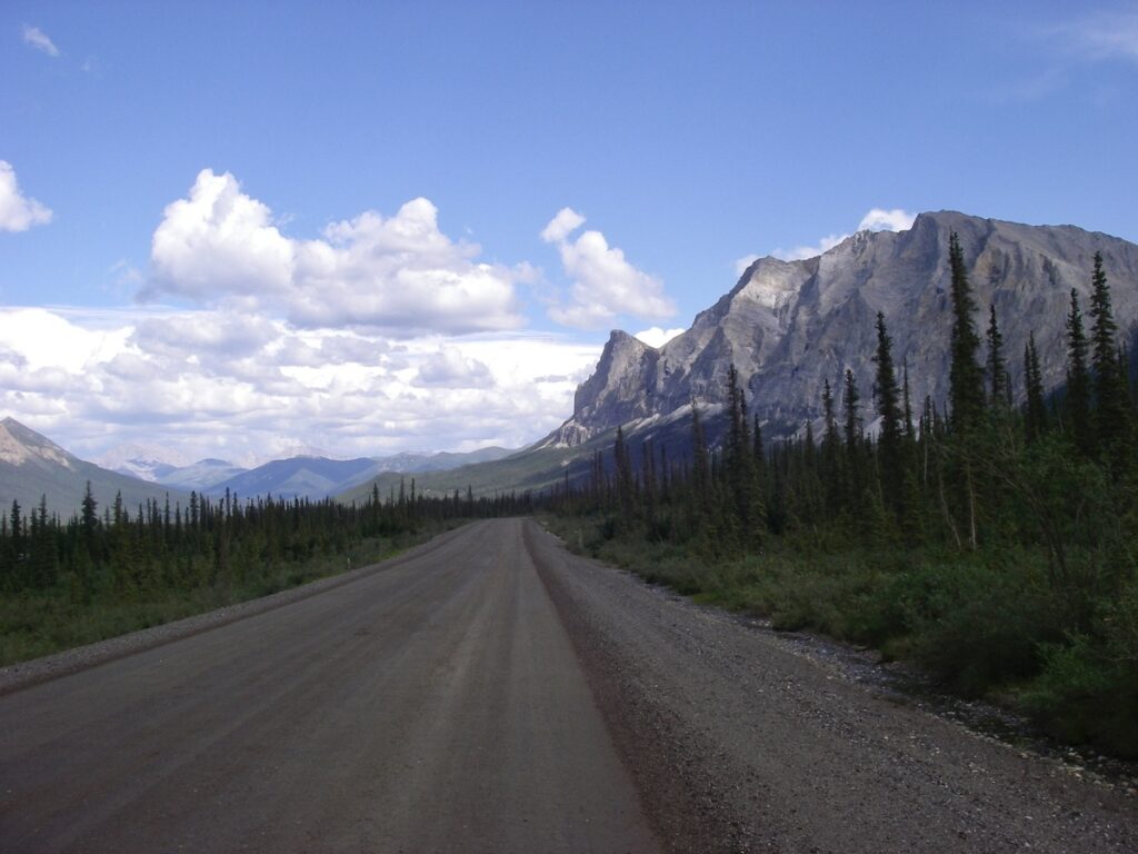 The Dalton Highway in Alaska, with a clear view leading straight to the rugged mountains in the distance, under a bright sky dotted with fluffy clouds, bordered by dense rows of evergreen trees.