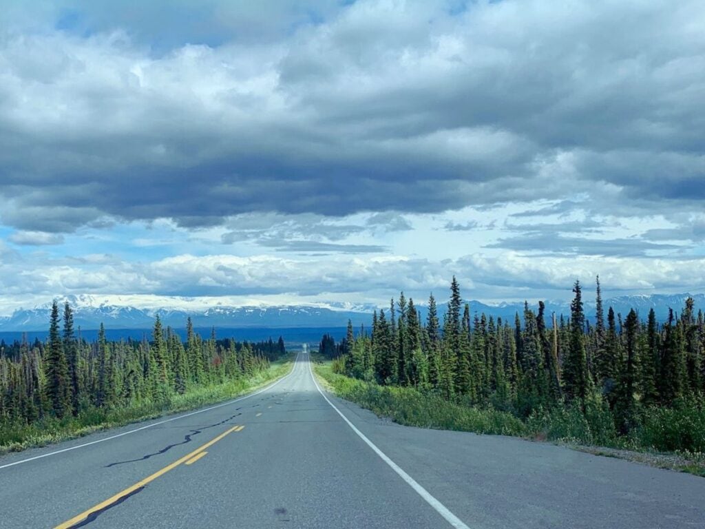 The Edgerton Highway in Alaska stretches forward, flanked by rows of tall, thin coniferous trees with snow-covered mountains on the horizon under a dramatic cloudy sky.
