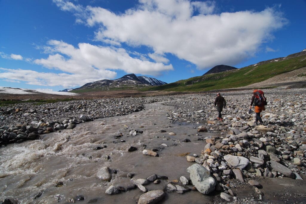 My husband and I with backpacks crossing a rocky glacial stream with clear rushing waters, set against a backdrop of green slopes and snow-capped mountains under a blue sky dotted with white clouds.