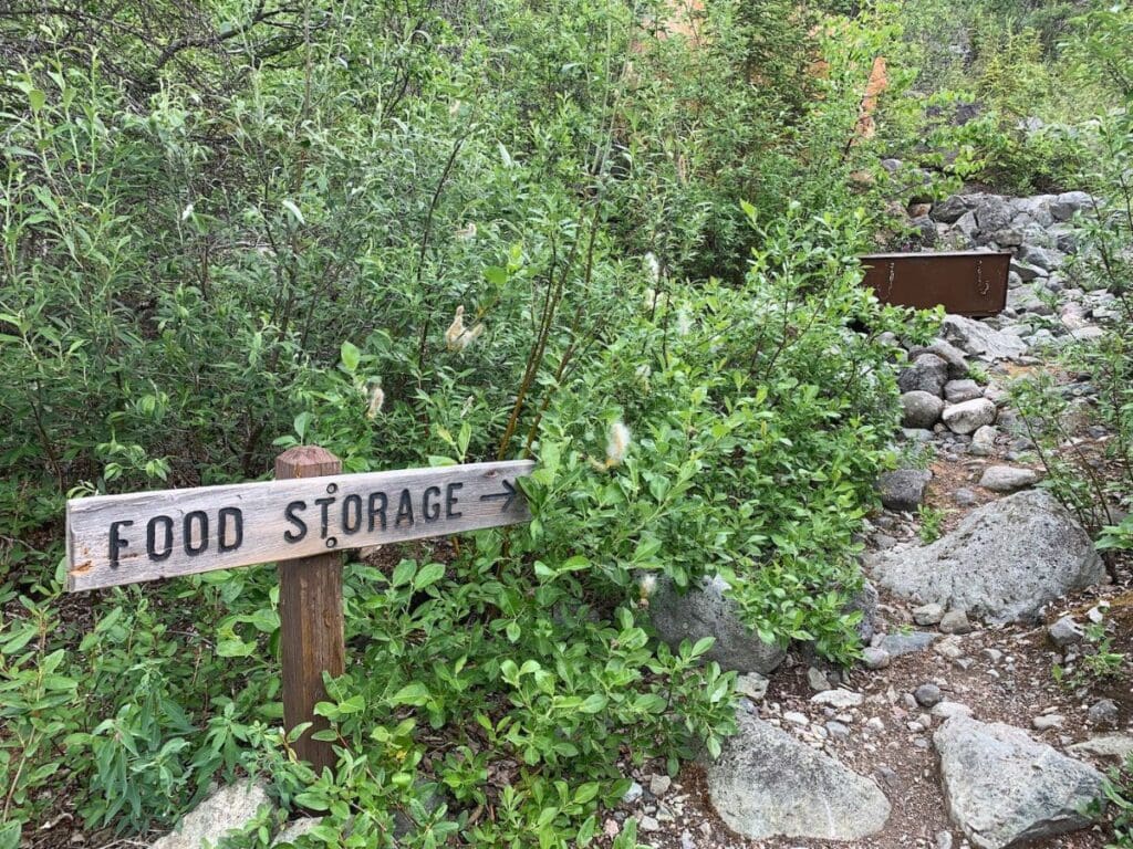 A wooden sign reading 'Food Storage' with an arrow pointing to the right, situated among lush green foliage and a rocky path, indicating the direction to a campsite's food storage area to protect against wildlife.