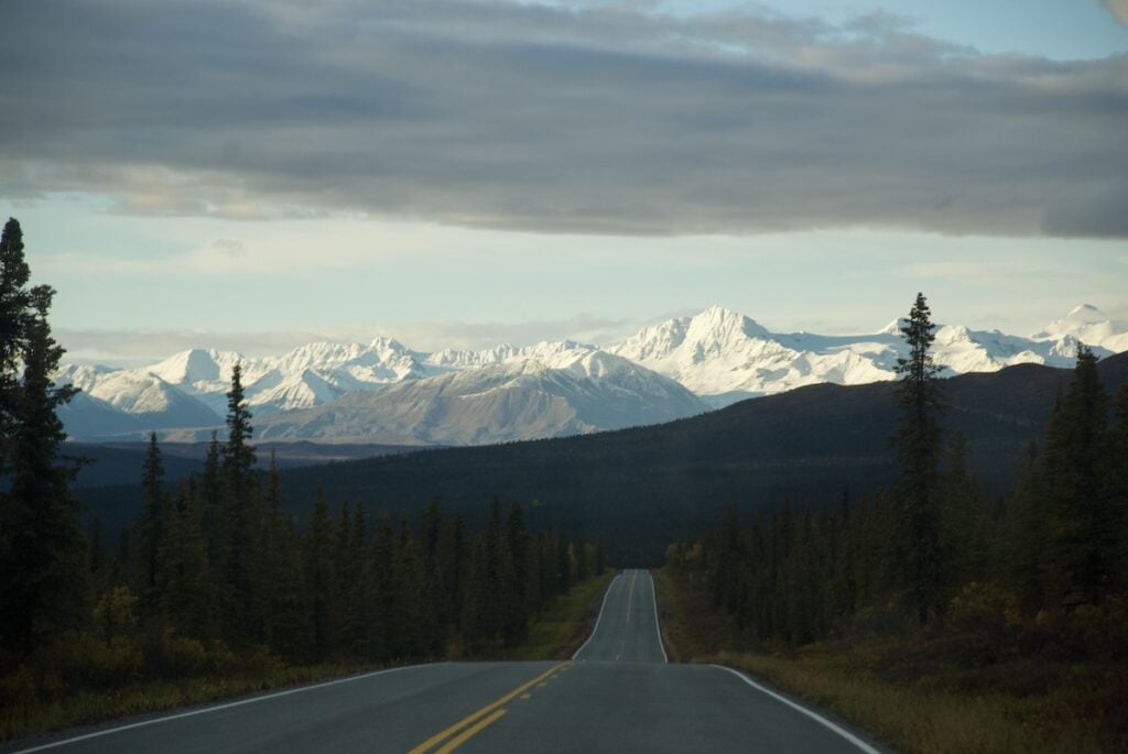 View from the Richardson Highway in Alaska, with the road stretching towards snow-capped mountains in the distance, flanked by evergreen forests under a partly cloudy sky.