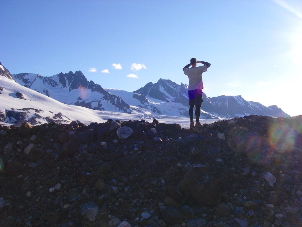 Silhouette of my husband, Matt, standing atop a rocky terrain gazing towards distant snowy peaks under a clear blue sky in Alaska, with the sun casting a lens flare.