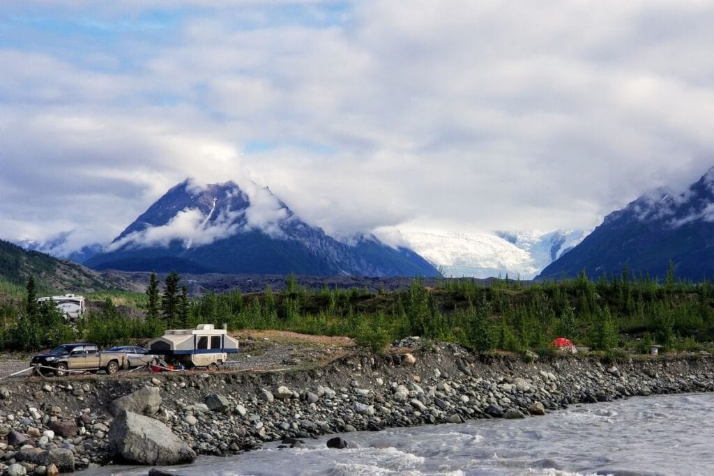 Base camp setup with a camper and vehicles by the rocky banks of a river near Kennecott, Alaska, with majestic, cloud-veiled mountains and the distant Kennicott Glacier providing a stunning backdrop, reflecting the rugged beauty of Alaskan wilderness camping.