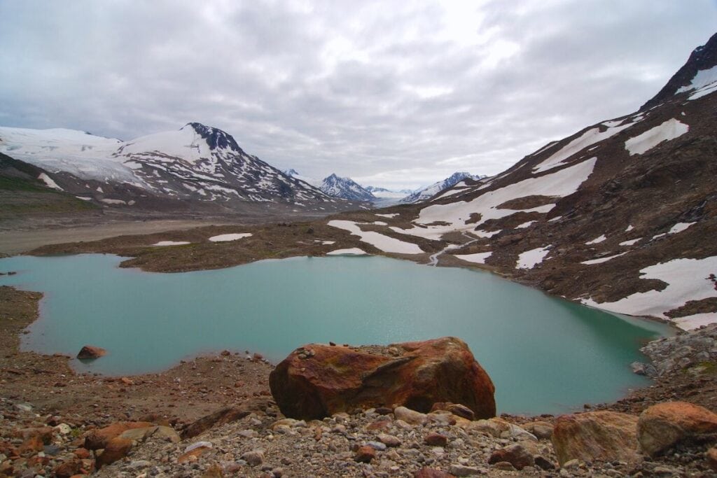 A serene glacial meltwater lake in hues of turquoise, nestled among the brown and snow-patched slopes of Alaska, with rugged mountains and overcast skies in the distance.