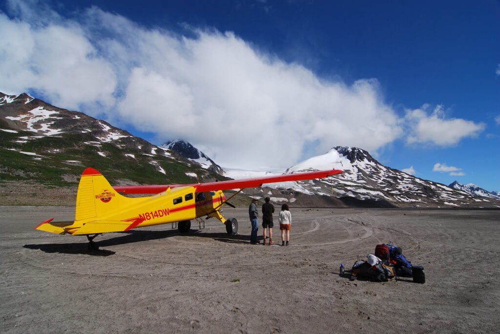 A bright yellow bush plane with red accents parked on a vast, sandy Alaskan landscape, with a small group of backpackers gathered nearby, preparing for an adventure with snow-capped mountains and blue skies in the background.