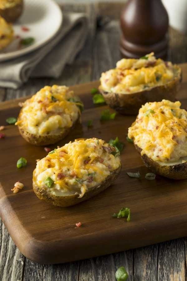 Grilled twice baked potatoes sit on a cutting board. They are topped with cheese and bacon bits. These are such a great unique camping food idea.