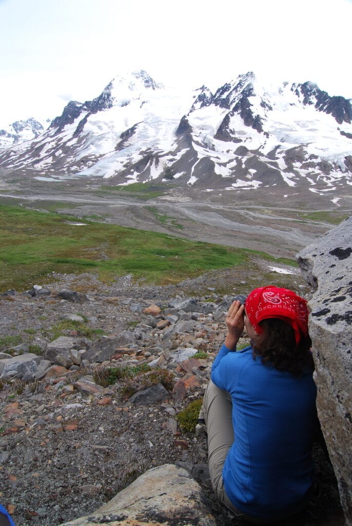 This is me, Heather ,wearing a red bandana and blue shirt gazes at the majestic snow-covered peaks in Wrangell-St. Elias National Park, Alaska, seated among rocky terrain with lush green valleys below.