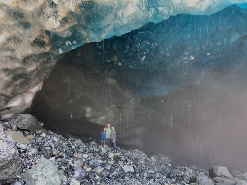 My kids and I are standing inside an ice cave near Kennecott, with a majestic blue ice ceiling above and rocky ground beneath, accentuated by the natural light filtering through the glacier.