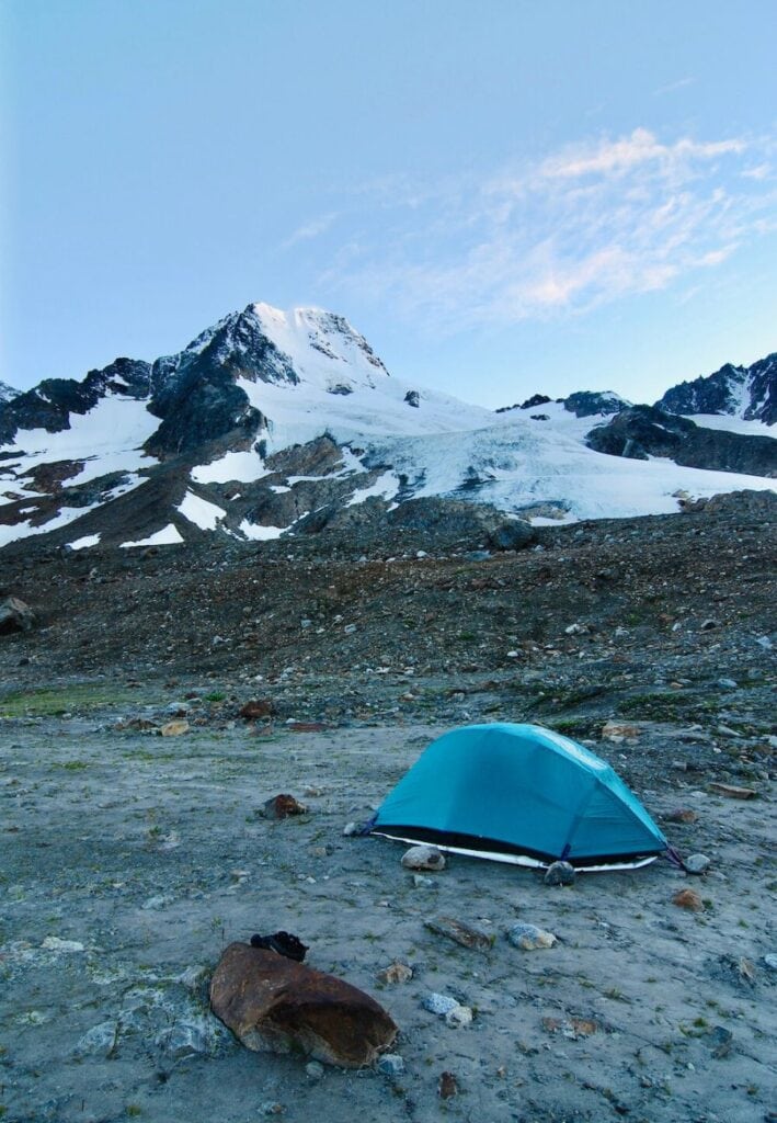 A solitary blue tent on a barren, rocky terrain at the base of a majestic snow-capped mountain peak, with the early morning sky beginning to light up in Wrangell St. Elias National Park.
