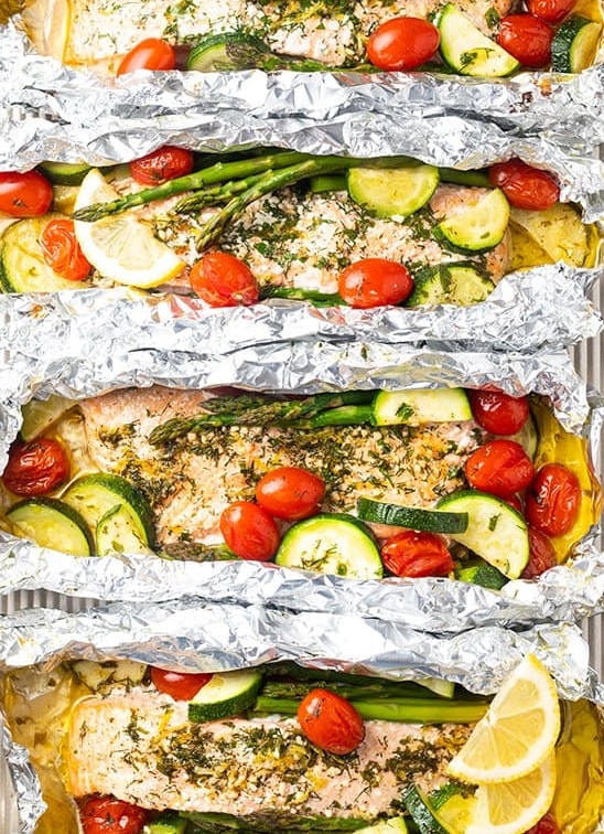 Baked salmon foil packets are a colorful and yummy idea for camping as they are prepared with asparagus, zucchini, and tomatoes.