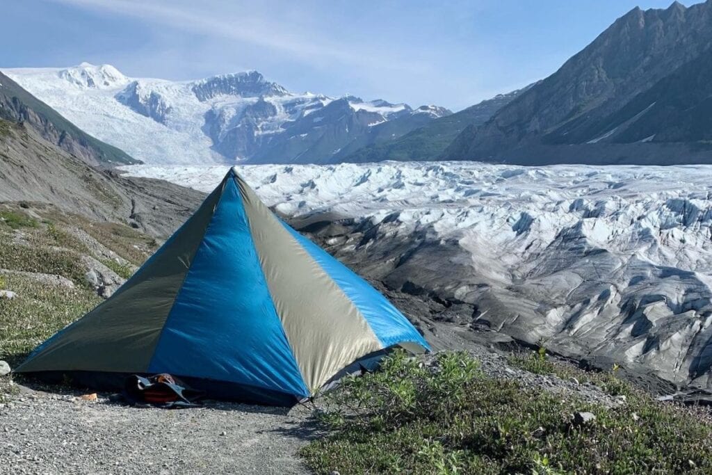 A two-tone blue and grey tent pitched on a rocky terrain with a stunning view of the sprawling Root Glacier and rugged mountains under a clear blue sky.