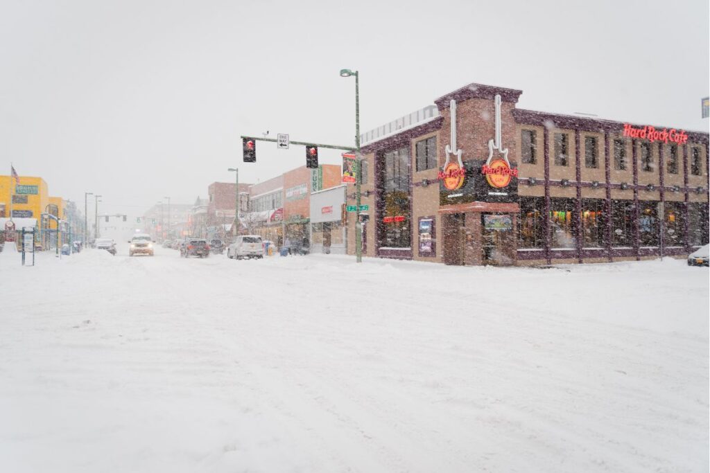 A snowy day in downtown Anchorage with vehicles cautiously navigating the snow-covered roads; the Hard Rock Cafe stands out with its signature guitar sign amidst the whiteout conditions.