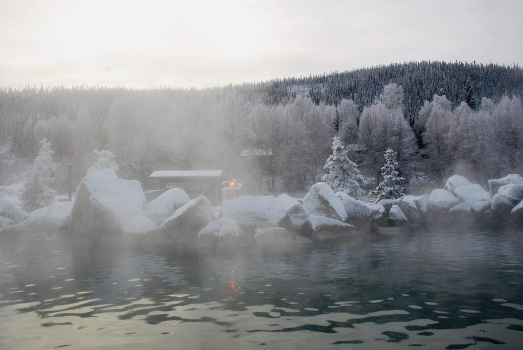 Steam rises from the warm waters of Chena Hot Springs in Alaska on a chilly day, with snow-covered rocks in the foreground and frosted trees against a forested hill in the background during the soft light of November.