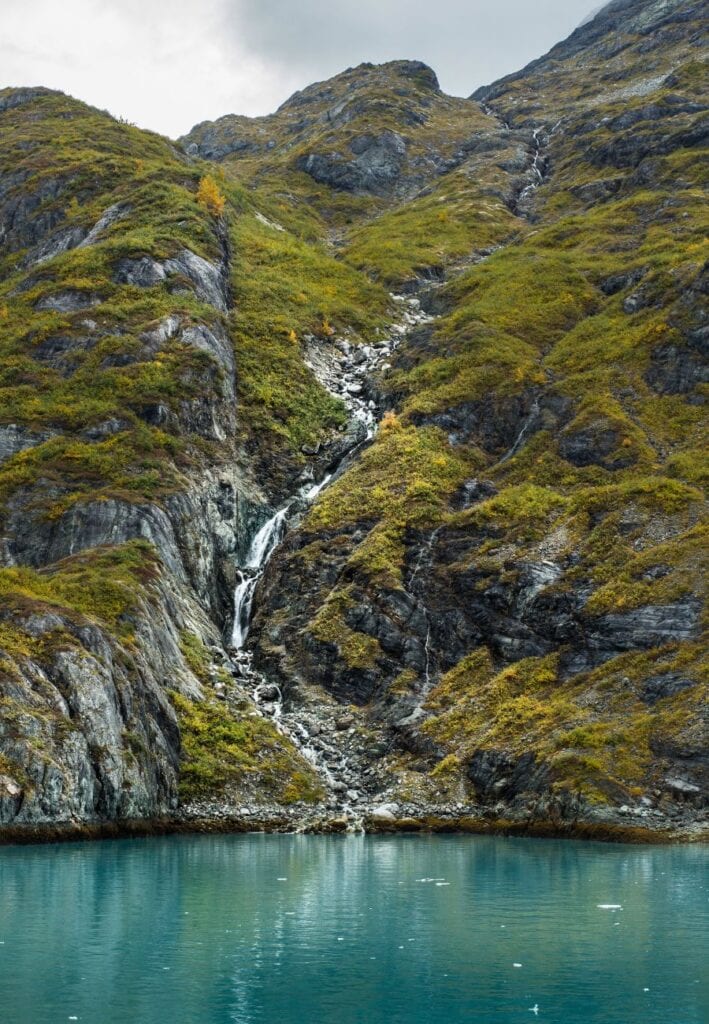 Glacier Bay, Alaska, in November, showcasing a serene landscape where towering mountains tinted with warm green and a waterfall meet the still, reflective green-blue waters. 