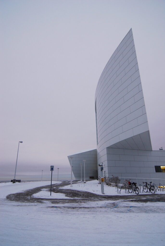 A modern, curved architecture of the Museum of the North in Alaska, with a light dusting of snow on the ground, and a bike rack with several bicycles in the foreground under a muted winter sky.