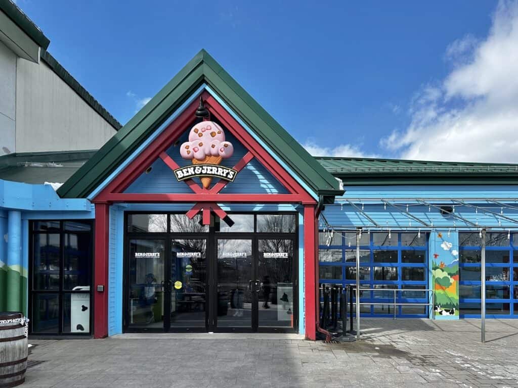The entrance to Ben and Jerry's in Waterbury, Vermont is cheerily painted birght light blue, red and green. There are blue garage-style doors with windows and a cute mural of the countryside with a cow on one small wall.