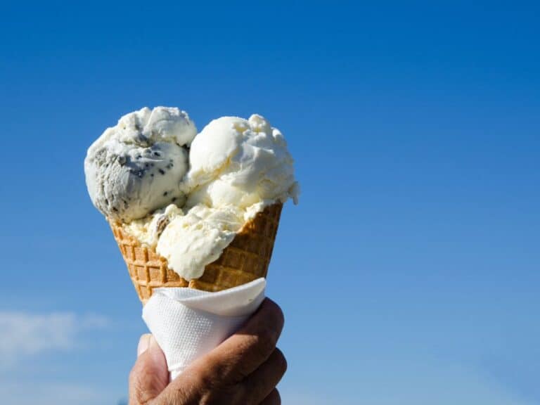 An ice cream waffle cone with several scoops is held against the blue sky.