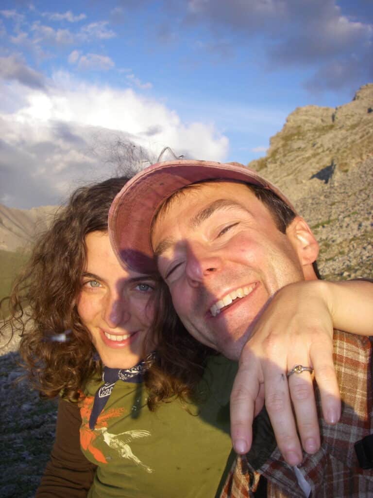 My husband, Matt, and I are illuminated by the midnight sun at 12 AM in the Arctic Circle, sharing a joyful embrace in a selfie. I have curly hair and wear a green shirt with a bird design, while my husband, on the right, dons a cap and plaid shirt. We are both smiling broadly, despite the presence of mosquitoes around.