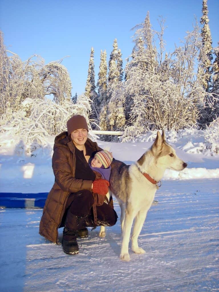 This is me holding my daughter, an infant at the time, with my Alaskan husky. We went out with my husband, Matt to harvest our Christmas tree. The snow trees behind me have beautiful snow on them reflecting in the brightest part of the day.