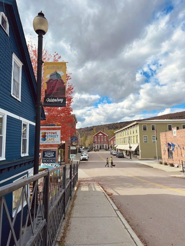 Downtown Waterbury has a cherry vibe with vibrant colors especially in the fall months. Looking down the street there is a couple walking a dog. In the foreground a bright blue building and store signs create contrast.