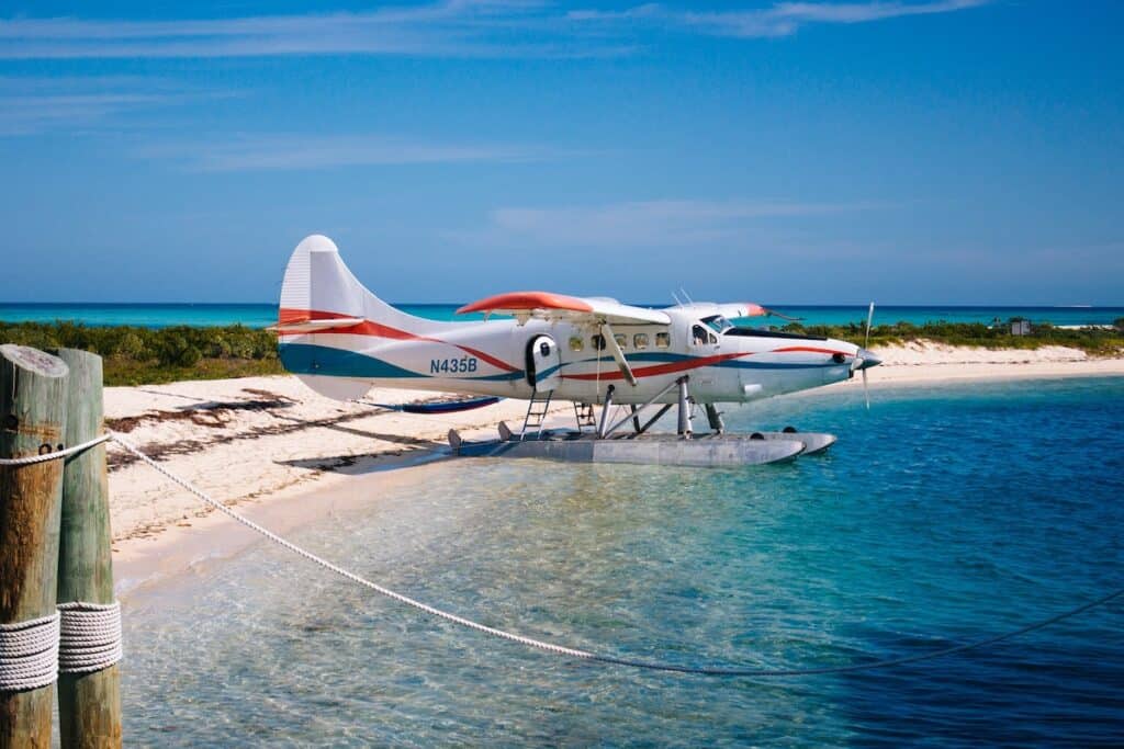 A white sea plane with blue and red stripes sits in the water at the edge of a strip of sandy land at Dry Tortugas National Park in Florida. The water is a deep turquoise. 
