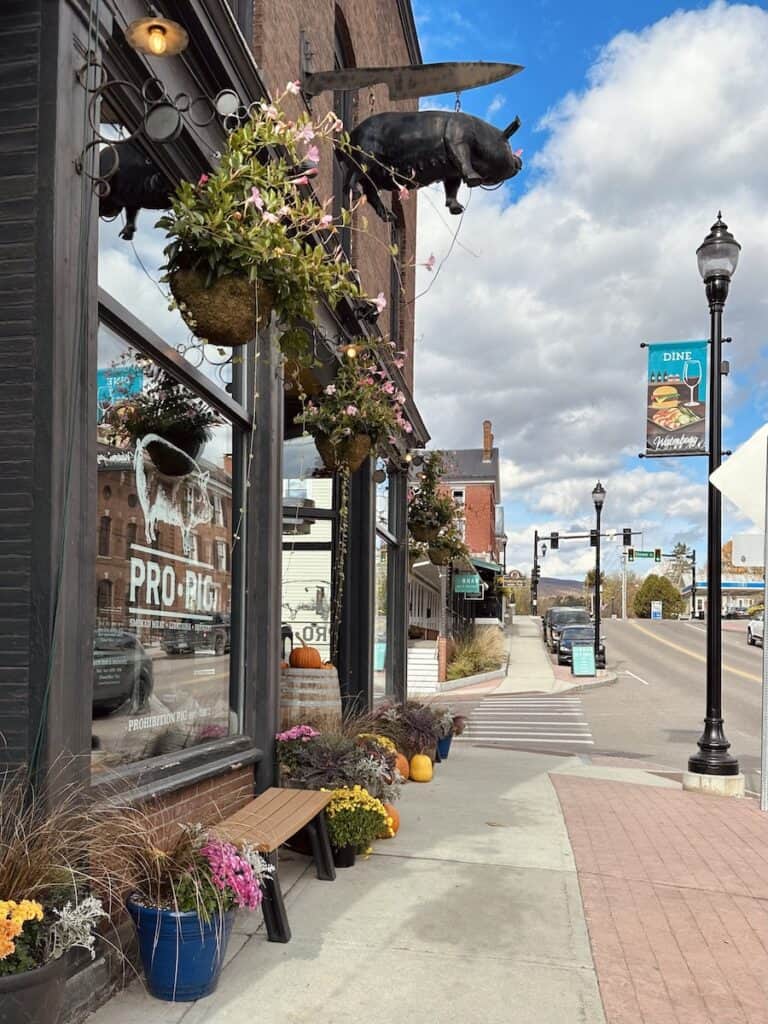 The exterior of Prohibition Pig is decorated with fall flowers and more. The view looks down the street in Waterbury on a sunny autumn day. Decorative black lamp posts line the block.
