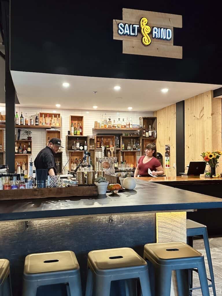 The interior of Salt and Rind. Owners Britt and Luke are in view. The dark stone bar counter contrasts against the metal bar stools in the foreground.