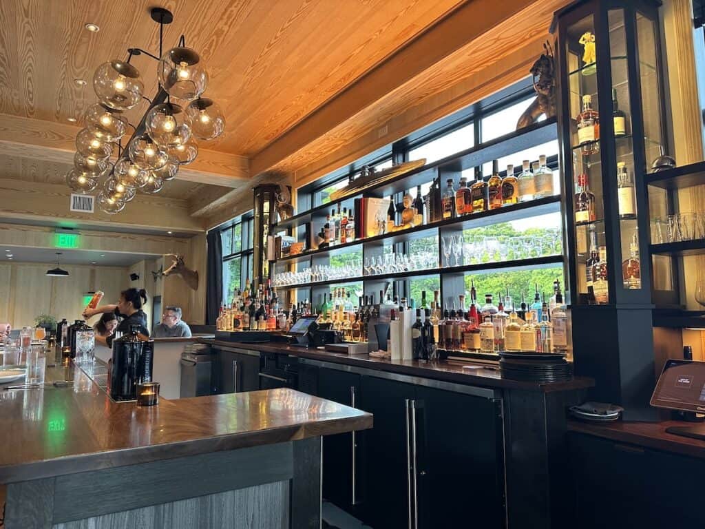 This is the bar at Hen of the Wood restaurant. It is modern and bright with windows backing the open shelving on which the bottles and glasses are stored. A bartender pours a drink.
