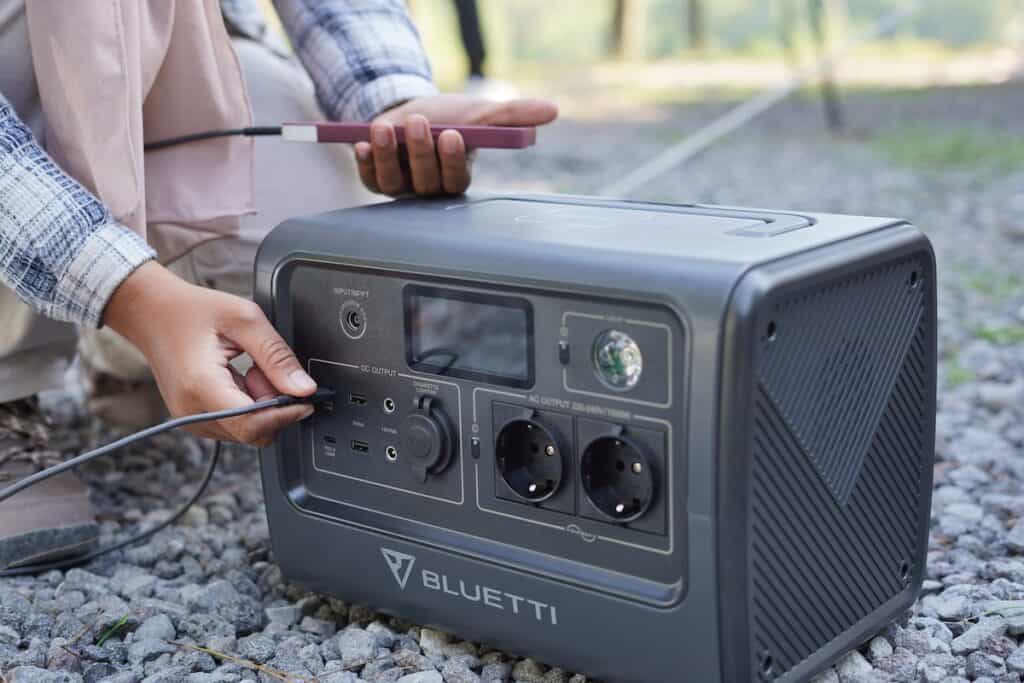 A Bluetti generator sits on the ground on gravel. A person holds a rose colored cell phone as they plug it into the generator to charge. It is a sunny day and the forest is seen in the background.