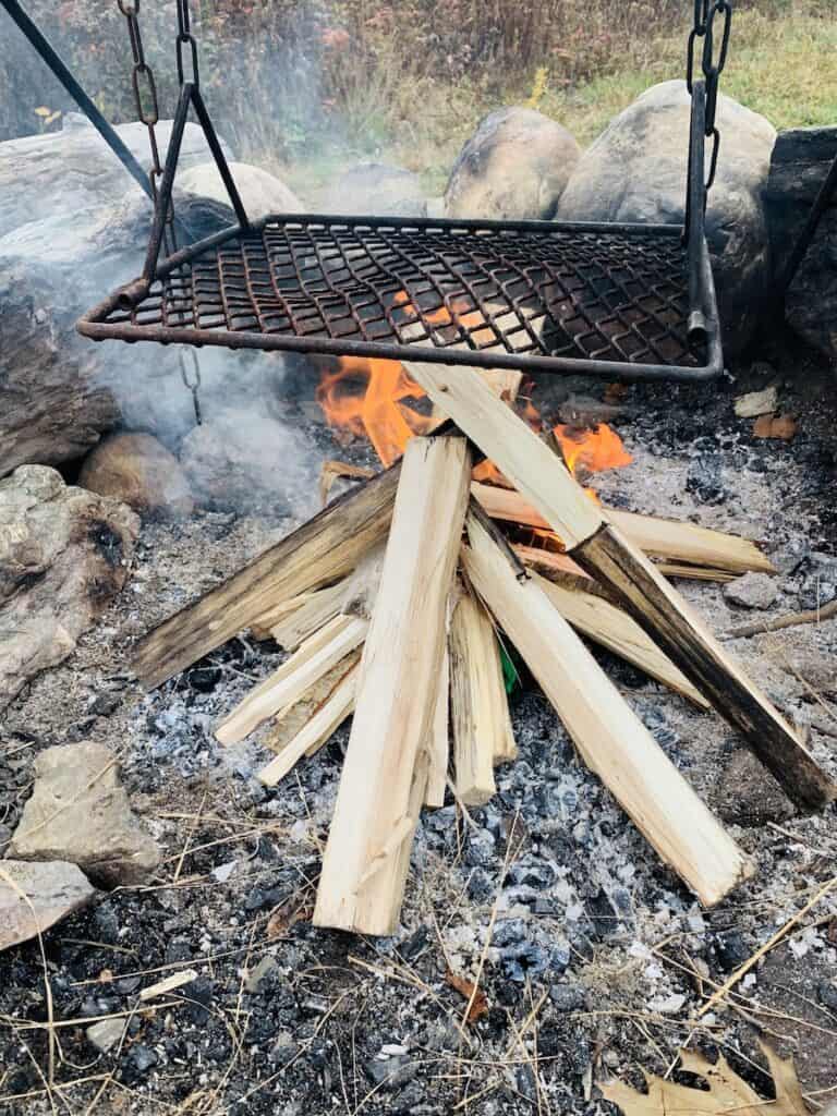 An iron grate hangs over a tripod of firewood that has just been lit for a campfire. Large stones line the fire circle border. 