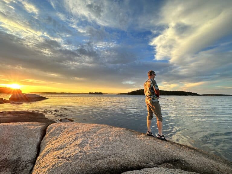My husband fishes while standing on the edge of the water on a rock. This is the ocean in Maine. A gorgeous sunset with golden light is in the backdrop and casts a glow on everything.