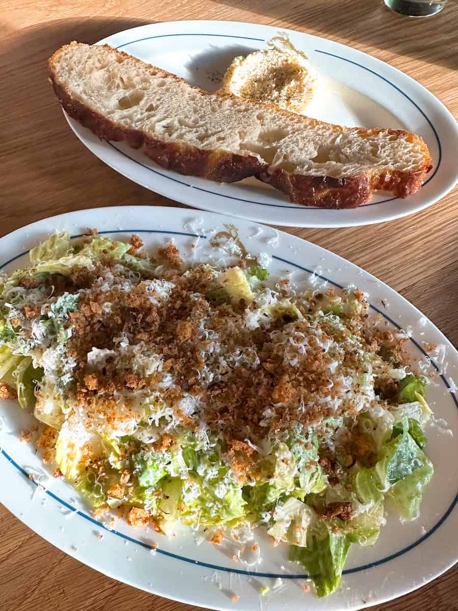 A thick slice of rustic bread served with a dollop of whipped butter sprinkled with cracked pepper sits on a white plate. In the foreground, a Caesar-style salad is topped generously with finely grated cheese and crispy breadcrumbs.