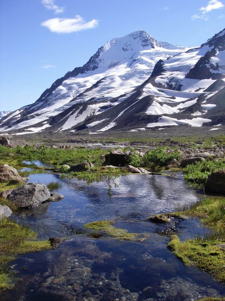 The backcountry near McCarthy Alaska is exquisite with snow capped mountains and glacier pools dappled with bits of green tundra. It is a sunny day with blue skies.