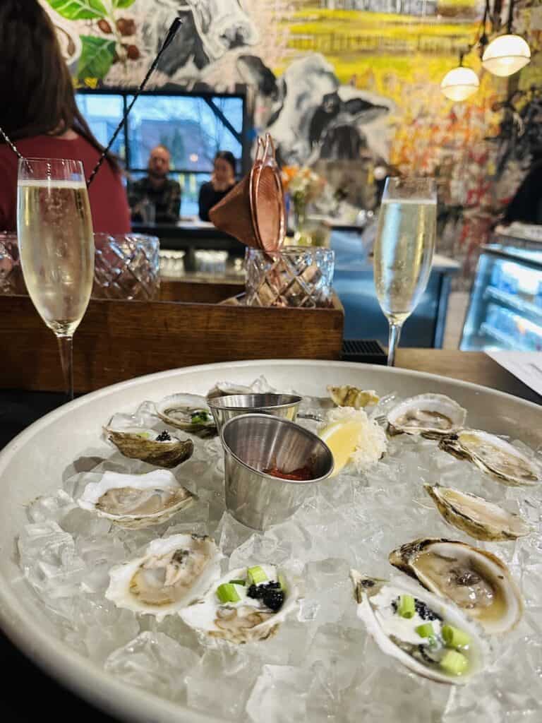 A selection of fresh oysters it on ice in a serving tray atop a bar. Two glasses of champagne are behind the platter and the bar scene with a vibrant mural wall is in the background.