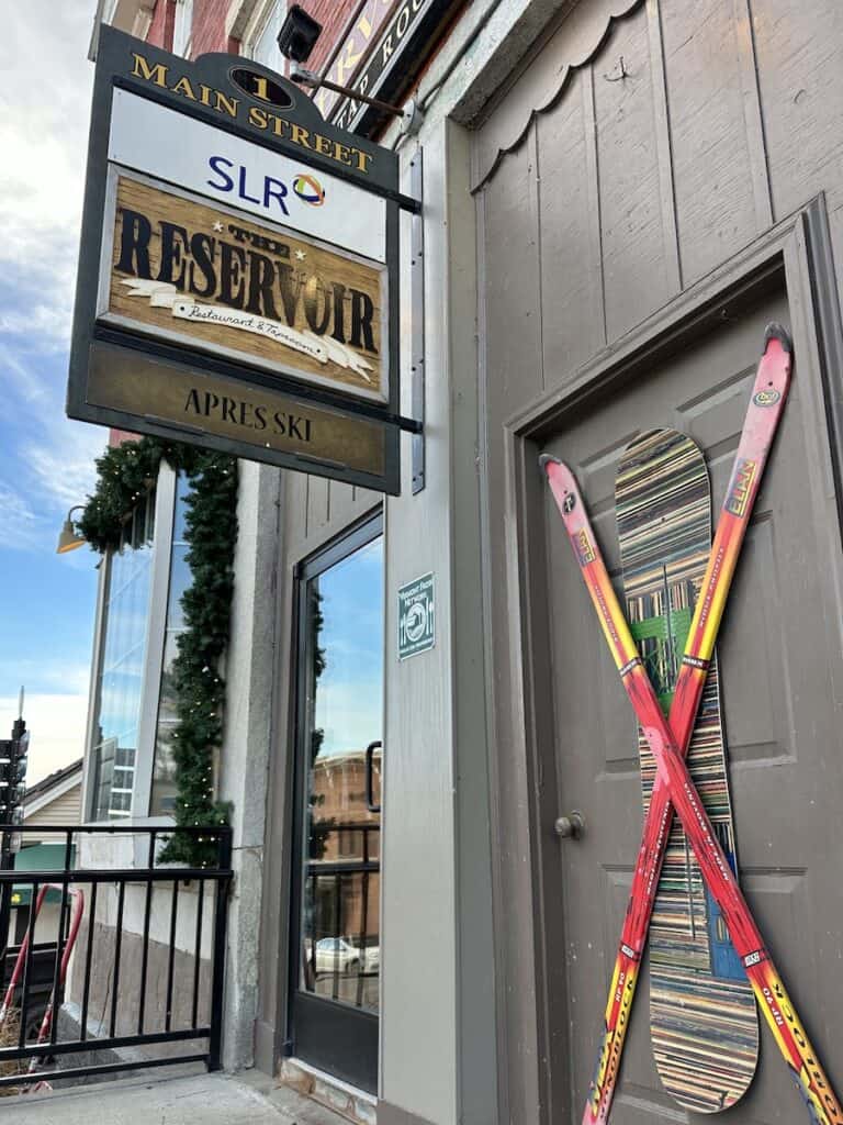 The exterior of The Reservoir sports bright red and yellow skis that hang crossed over a striped snowboard on the door next to the entrance. The holiday greenery and lights hanging can be seen next to the entrance door as well.