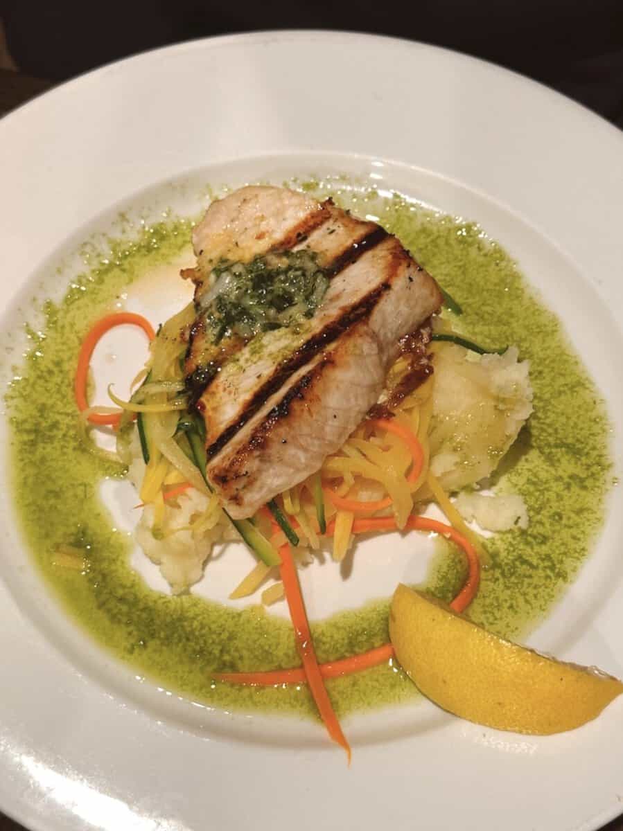 A swordfish dinner special at Fin and Fern has a fresh herb sauce lining the rim of the plate. Shredded sauteed vegetables, mashed potatoes, and a slice of lemon accompany the fish.