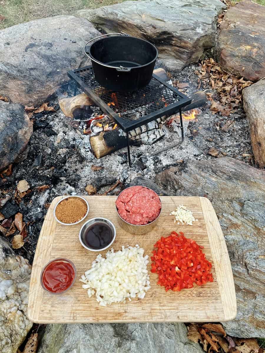 A cutting board sits on the rock border of the fire pit. On the cutting board are piles of ingredients such, as chopped red pepper, minced onion, and ground beef. In little bowls are ketchup, Worcestershire sauce and dried ingredients like sugar and salt. A Dutch oven sits above the campfire on metal grate.